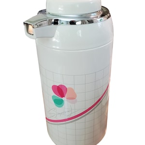 May include: White insulated beverage dispenser with a silver spout and accents. The dispenser has a white lid and a floral design with the word "Symphony" in cursive. The body of the dispenser has a grid pattern.