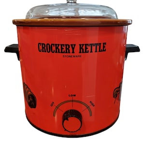 May include: Red Crockery Kettle with the words "Crockery Kettle" in black lettering. The slow cooker has a glass lid with a decorative handle, a wooden rim, and black handles on the sides. The control knob has settings for low and high.