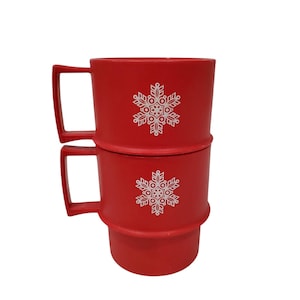May include: Two stackable red plastic mugs featuring a white snowflake graphic on the front. Each mug has a square-shaped handle and a ridged design that allows them to fit securely one on top of the other for compact storage and organization.