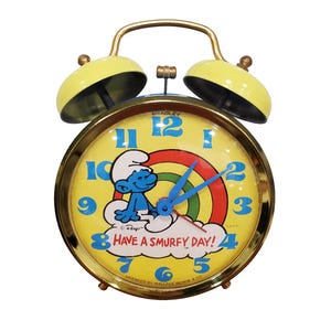 May include: A vintage yellow and gold alarm clock featuring a Smurf character on the clock face. The clock face has blue numbers, blue hands, and the text "HAVE A SMURFY DAY!". The clock has two yellow bells and a gold handle.