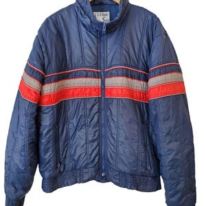 May include: A vintage navy blue Alpine Ski jacket with red and gray stripes across the chest. The jacket has a high collar, a full zipper, and ribbed cuffs and hem. The jacket is hanging on a wooden hanger.