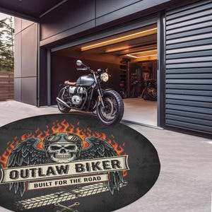 May include: A round black rug with a skull wearing a helmet and wings, the text "OUTLAW BIKER" and "BUILT FOR THE ROAD" is displayed. A motorcycle is parked in front of a garage.