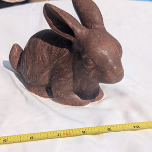 May include: A brown ceramic rabbit figurine in a seated position. The rabbit has long ears and is resting on its front paws. A yellow measuring tape is in the foreground. The figurine is approximately 8 inches long.