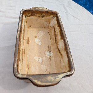 May include: Rectangular ceramic loaf pan with a light beige interior and a brown and gray rim. The pan has two small handles on the sides. The interior has white spots and brown streaks.