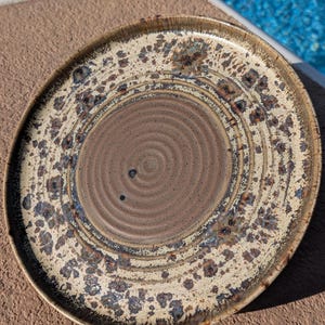 May include: A round ceramic plate with a spiral design in the center. The plate has a beige background with brown and blue speckled accents. The center is a darker brown with a spiral pattern.