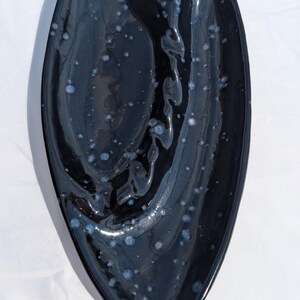 Könnte beinhalten: Eine schwarze Keramik-Servierschale mit einer einzigartigen, länglichen Form. Die Schale hat eine glänzende Oberfläche mit gesprenkelten blauen und weißen Akzenten. Das Design weist eine zentrale Vertiefung und einen geschwungenen Rand auf.