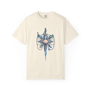 Peut inclure: T-shirt crème avec une illustration détaillée d'un motif en forme d'épée. Le motif comprend des pierres précieuses bleues, un coquillage et des chaînes. L'œuvre d'art a un thème fantastique ou nautique.