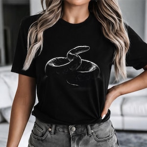 May include: Black t-shirt featuring a grayscale illustration of a coiled snake. The snake design is centered on the shirt. The shirt is paired with gray denim shorts. The overall aesthetic is casual and modern.