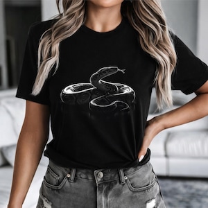 Może przedstawiać: Czarny t-shirt z szczegółową, szarą ilustracją zwiniętego węża. Wzór węża jest wyśrodkowany na koszulce, tworząc uderzający kontrast z ciemną tkaniną. Koszulka ma klasyczny okrągły dekolt.