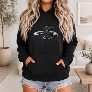 Peut inclure: Sweat à capuche noir avec une illustration en niveaux de gris d'un serpent enroulé. Le motif détaillé est centré sur le devant. Le sweat-shirt a une poche kangourou. La personne porte un short en jean.