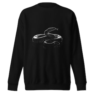 Puede incluir: Sudadera negra con una imagen en escala de grises de una serpiente enroscada con la lengua fuera. El diseño está centrado en la parte delantera de la sudadera, sobre un fondo oscuro. La imagen en tonos grises crea un contraste llamativo.