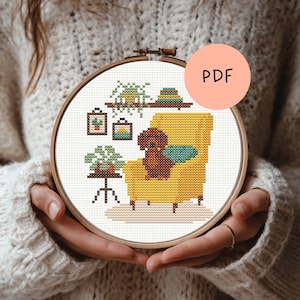 Op de afbeelding: Een borduurring met kruissteek met een gezellige woonkamerscène. Het ontwerp omvat een gele fauteuil, een bruine hond, potplanten en ingelijste kunstwerken. Een perzikkleurige cirkel met de tekst "PDF" staat rechtsboven.