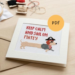 Op de afbeelding: Kruissteekkaart met een teckel piratenontwerp. De tekst "Keep Calm and Sail On Matey" is rood. De teckel draagt een piratenhoed en jas. De achtergrond is lichtbeige.