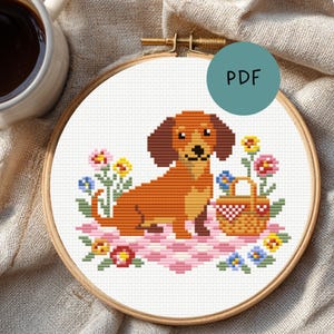 Op de afbeelding: Kruissteekborduurwerk in een houten hoepel met een teckel, een picknickmand en bloemen. De hond is bruin en bruingeel, op een roze en wit geblokt kleed. Het woord "PDF" staat in een turquoise cirkel.