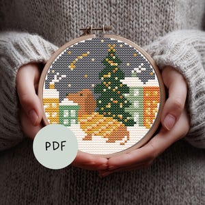 Op de afbeelding: Een borduurring met kruissteek met een wintertafereel met een teckel, een kerstboom en met sneeuw bedekte gebouwen. Het ontwerp gebruikt tinten bruin, groen, geel en wit tegen een donkergrijze achtergrond. Het woord "PDF" is zichtbaar op een lichtgroene cirkel.