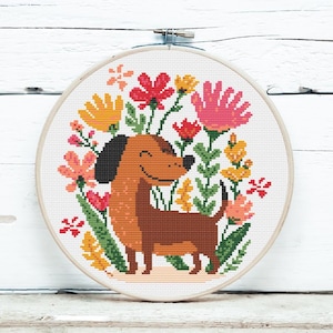 Op de afbeelding: Een borduurring met kruissteek met een lachende teckel omringd door kleurrijke bloemen. De hond is bruin met een zwart oor en de bloemen zijn roze, geel en rood. De ring staat tegen een witte houten achtergrond.