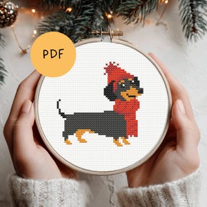 Op de afbeelding: Een kruissteekpatroon van een teckel met een rode muts en sjaal, weergegeven in een houten borduurring. De hond is zwart, bruin en rood, met de tekst "PDF" in een gele cirkel.