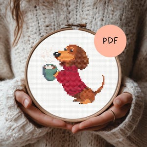 Op de afbeelding: Een kruissteek borduurring met een teckel die een rode trui draagt en een turquoise mok met stoom vasthoudt. De hond is bruin en bruin. De ring heeft een houten frame en het woord "PDF" staat op een roze cirkel.