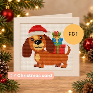 Op de afbeelding: Een vierkante wenskaart met een kruissteekpatroon van een bruine teckel met een rood-witte kerstmuts, die gestapelde kleurrijke cadeautjes op zijn rug draagt. De kaart bevat de tekst PDF en DIY Christmas card.