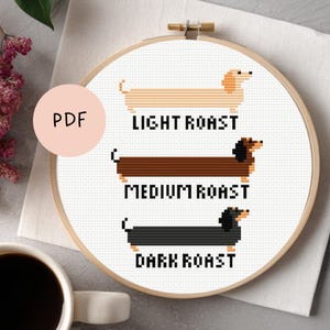 Op de afbeelding: Een kruissteekpatroon in een houten borduurring toont drie teckels, die elk een koffiebranding vertegenwoordigen: licht, medium en donker. De teckels zijn in tinten beige, bruin en zwart. De tekst "PDF" staat in een roze cirkel.