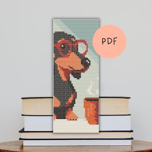 Op de afbeelding: Een kruissteek bladwijzer met een teckel met een rode bril, die een koffiekopje vasthoudt. De bladwijzer heeft een lichtblauwe achtergrond en is gemarkeerd met "PDF" in een perzikkleurige cirkel. De bladwijzer ligt op een stapel boeken.