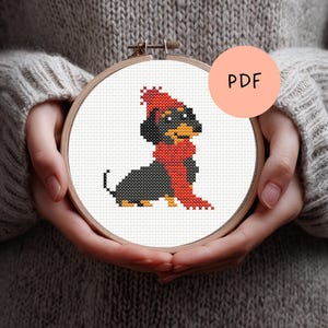 Op de afbeelding: Een kruissteekborduurwerk van een teckel met een rode hoed en sjaal, gehouden in een houten hoepel. De hond is zwart en bruin, met het woord "PDF" in een perzikkleurige cirkel op de achtergrond.