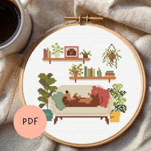 Op de afbeelding: Kruissteekpatroon in een houten hoepel met een gezellige woonkamerscène. Het ontwerp toont een bank met een hond, boekenplanken, planten en de tekst "PDF" op een perzikkleurige cirkel. Een kopje koffie staat in de linkerbovenhoek.