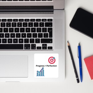 May include: A white sticker with the text "Progress > Perfection" and a target icon with a blue arrow. Below the text is a blue and purple graph. The sticker is on a laptop.
