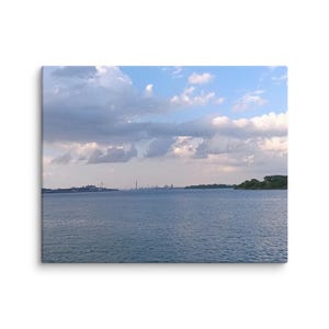 May include: A canvas print of a serene waterscape under a cloudy sky. The water is a deep blue, reflecting the sky's hues. Distant structures line the horizon, with a patch of green trees on the right.