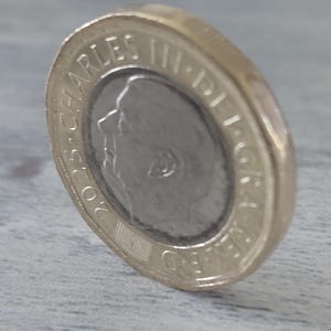 May include: A two-tone £2 coin, with a silver central disc and a gold-coloured outer ring. The coin shows a profile of a person and the text "2023 CHARLES III DEI GRA". The coin is tilted, showing its edge.