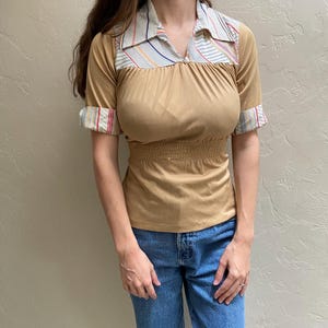May include: Tan short-sleeve top with a striped collar and cuffs. The top has a gathered waist and a subtle sheen. The collar and cuffs feature a pattern of thin stripes in red, blue, and yellow.