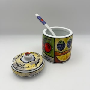 May include: A ceramic jam jar with a lid and spoon. The jar is decorated with images of cherries, blackberries, and a lemon slice. The lid has the word "Confiture" and "Jam" written on it. The spoon has a red strawberry design.