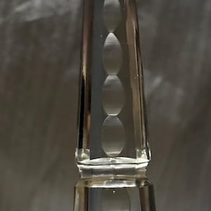 Könnte beinhalten: Ein klarer Kristallprismenanhänger mit facettiertem Boden und einer Reihe ovaler Vertiefungen. Ein silberfarbener Draht ist oben befestigt. Der Anhänger ist so konzipiert, dass er Licht reflektiert.