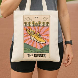 May include: Cream-colored tote bag featuring a tarot card design. The card depicts a yellow running shoe with wings, set against a backdrop of sunflowers, mountains, and a radiating sunburst. The text "THE RUNNER" is at the bottom.