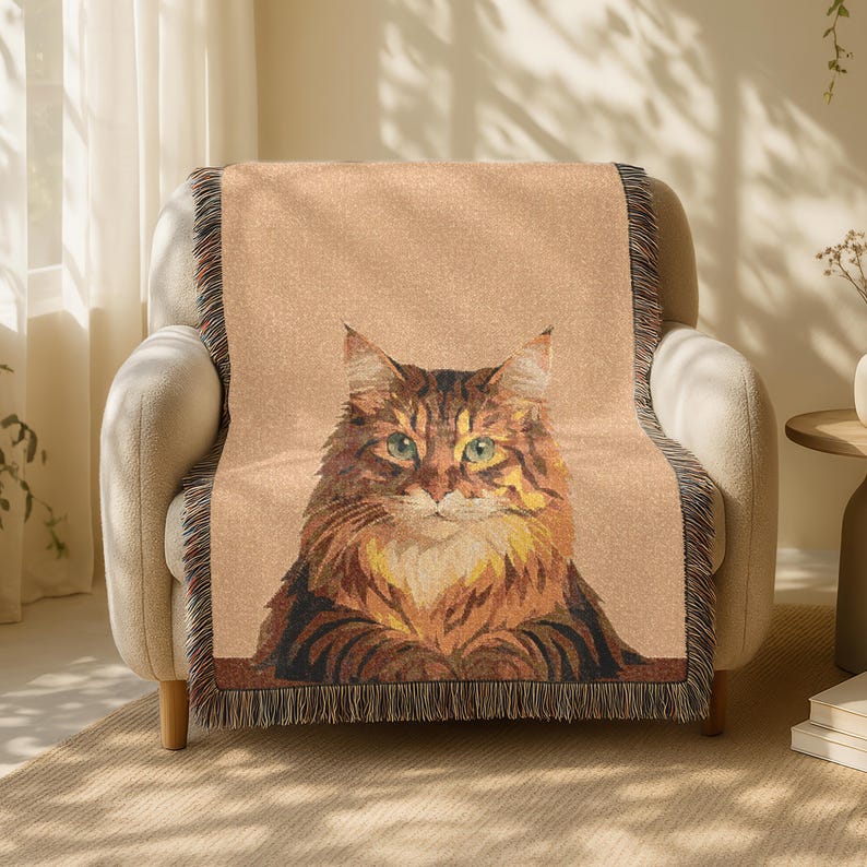 May include: A tan throw blanket featuring a detailed illustration of a cat with brown and orange fur and green eyes. The blanket has a fringed edge and is draped over a beige armchair.