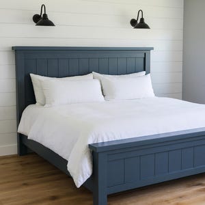 以下が含まれることがあります： ネイビーブルーの木製ベッドフレーム、ヘッドボードとフットボード付き、白い寝具と枕が組み合わされています。 ベッドは白い羽目板の壁に設置され、ヘッドボードの上には2つの黒い燭台があります。 部屋には木製の床があります。