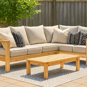 Op de afbeelding: Hoekbank en salontafel voor buiten. De bank heeft een licht houten frame met beige kussens en decoratieve kussens met zwart-witte geometrische patronen. De salontafel is gemaakt van bijpassend licht hout.