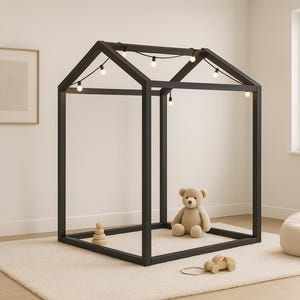 Puede incluir: Una estructura de cama con forma de casa de madera negra con luces de cadena, un oso de peluche beige y un pequeño coche de juguete de madera. La estructura de la cama está sobre una alfombra color crema, con un puf blanco y una planta en maceta cerca.