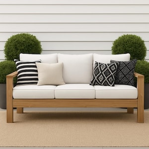 May include: A wooden outdoor sofa with white cushions and decorative pillows. The pillows feature black and white geometric patterns and stripes. The sofa is surrounded by green plants and sits on a beige rug.