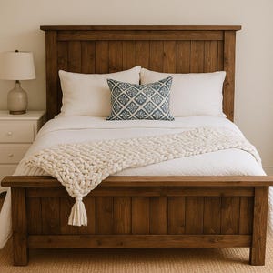 May include: A wooden bed frame with a headboard and footboard. The bed is made with a dark brown wood and has a white comforter, pillows, and a cream-colored knitted throw. A decorative blue and white patterned pillow is on top of the bed.