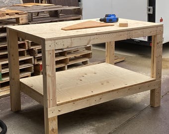 May include: A wooden workbench with a lower shelf and four sturdy legs. A blue power drill, a small wooden block, and a wooden template are on the top surface. The workbench is made of light-colored wood.