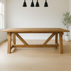 May include: A large, rectangular wooden dining table with a natural finish. The table has a sturdy base with angled supports and a thick tabletop. The room is well-lit, with a window and pendant lights visible.