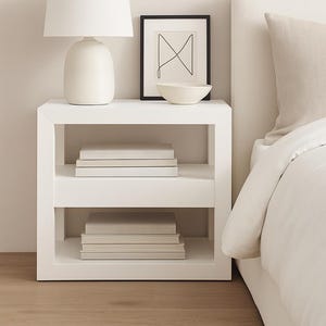 Può includere: Un comodino bianco con due ripiani, ciascuno con pile di libri. Una lampada bianca con base arrotondata è posta sopra, insieme a un'opera d'arte astratta incorniciata e una piccola ciotola bianca. Il tavolo è accanto a un letto con biancheria da letto bianca.
