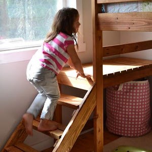 May include: A wooden bunk bed with a built-in ladder. A child in a pink and white striped shirt and gray pants is climbing the ladder. A pink polka-dot storage basket and a green pillow are visible.
