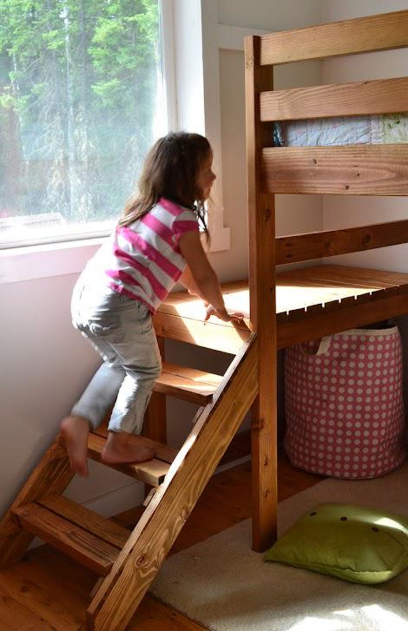 May include: A wooden bunk bed with a built-in ladder. A child in a pink and white striped shirt and gray pants is climbing the ladder. A pink polka-dot storage basket and a green pillow are visible.
