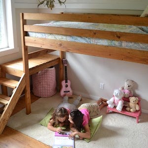 Peut inclure: Un lit mezzanine en bois avec un bureau et un escalier intégrés. Deux enfants sont allongés sur le sol, lisant un livre. Une guitare rose et un panier de rangement rose sont à proximité. Des ours en peluche sont assis sur un petit banc rose.