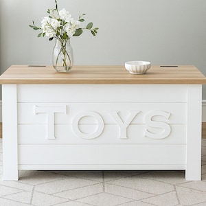 May include: A white toy chest with a light wood top. The word "TOYS" is displayed in large, white letters on the front. A vase of white flowers and a small white bowl sit on top of the chest.