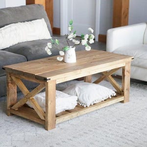 May include: A rectangular wooden coffee table with an X-shaped design on the sides. The table has a lower shelf holding white pillows. A vase of white flowers sits on top. The table is in a living room setting.
