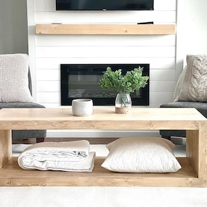 May include: A light wood rectangular coffee table with a lower shelf holding a folded blanket and a pillow. Two grey chairs with cable-knit pillows flank the table. A vase of greenery and a small ceramic pot sit on the table.