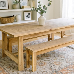 May include: A light-colored wooden dining table with a matching bench. The table has a rectangular top and sturdy legs. A decorative vase with greenery sits on the table. The bench is approximately 60 inches long.